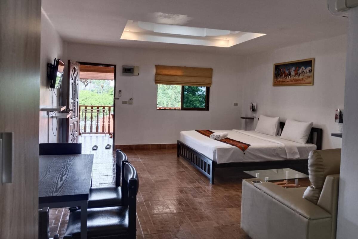 A room with a bed, a sofa, a dining table, and a TV. There is a window with a view of greenery outside.