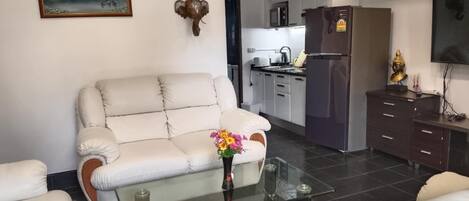 A living room with a sofa, a glass coffee table with flowers, a kitchen in the background, and a mounted TV.