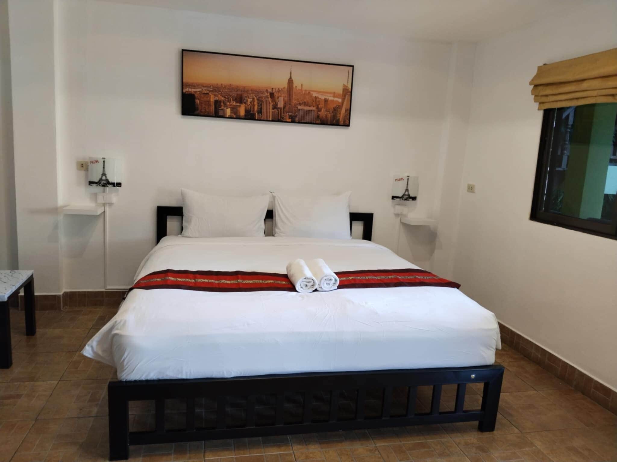 Habitación de hotel con una cama, un cuadro de un paisaje urbano, una mesita de noche, una lámpara y una ventana con vistas.