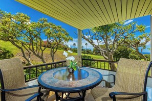Un espace extérieur aménagé, abrité et doté d’une table ronde et de deux chaises, offrant une vue sur un jardin luxuriant et sur l’océan.