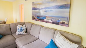 Ein Wohnzimmer mit einem beigefarbenen Ecksofa, einem Bild von Booten auf einem See und einer Tür zu einem anderen Raum.