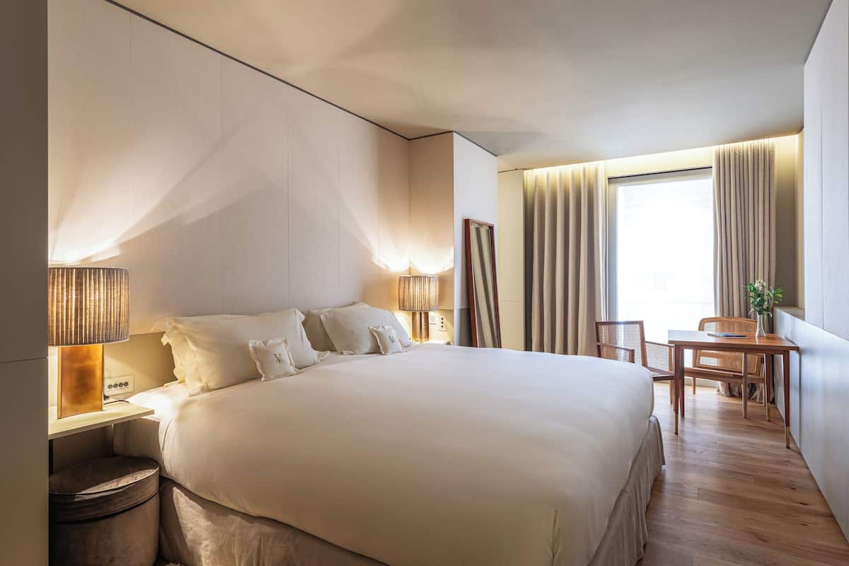 Ein modernes Hotelzimmer mit einem großen Bett, Nachttischlampen, einem kleinen Tisch mit Stühlen und Blick ins Freie durch Vorhänge.