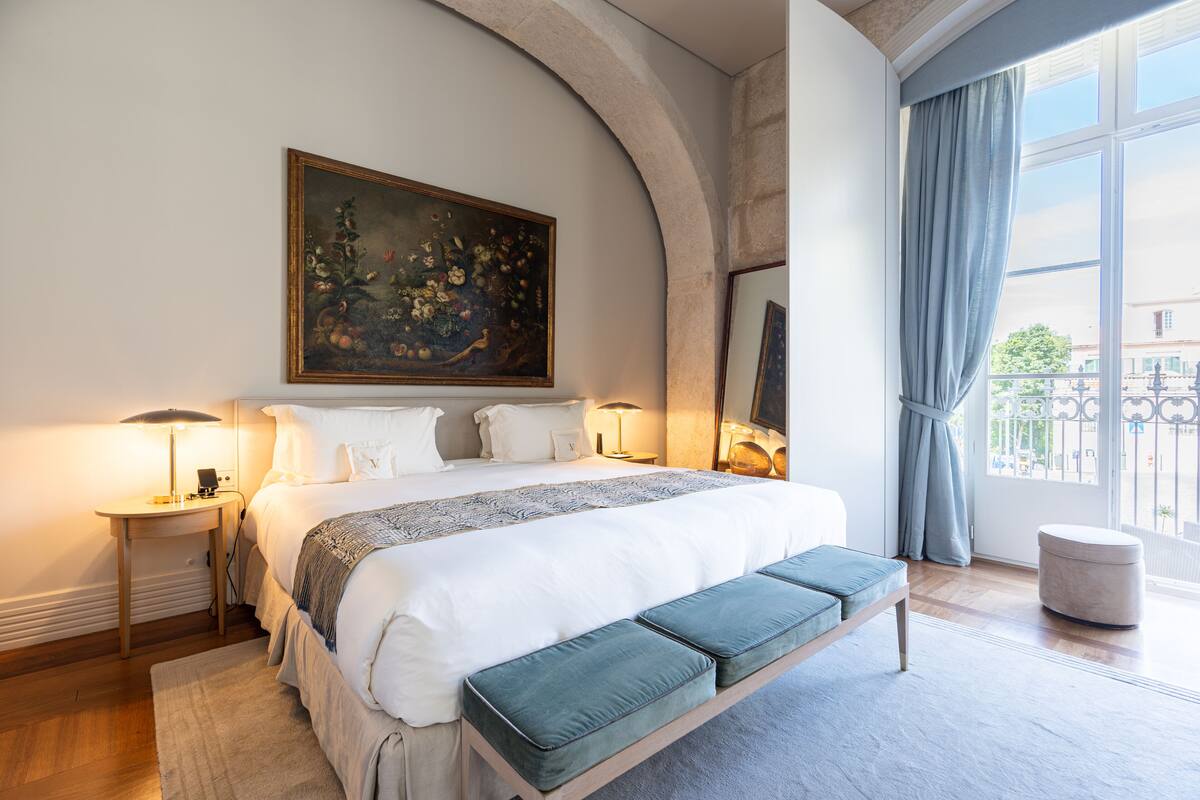 Ein Schlafzimmer mit einem großen Bett, einer Bank, einem Gemälde und einem Fenster mit Vorhängen.