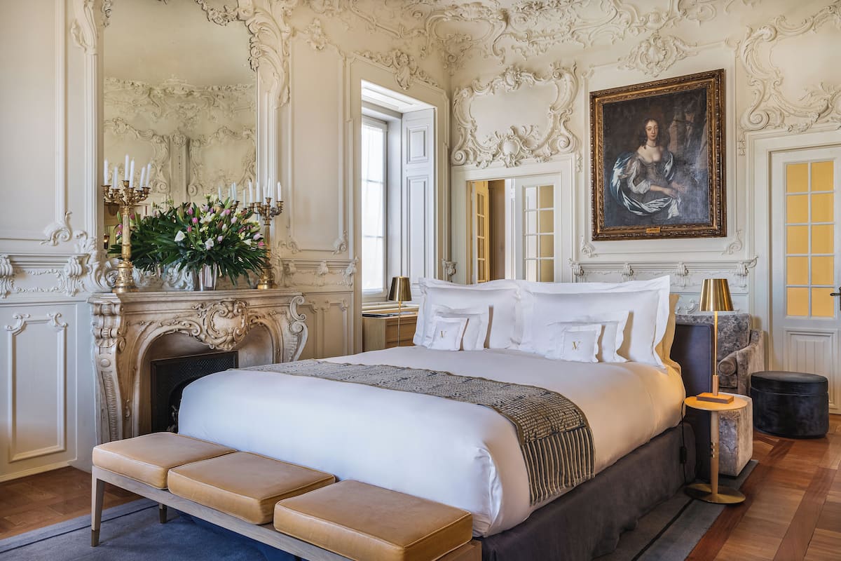 Ein luxuriöses Schlafzimmer mit einem aufwendigen Kamin, einem großen Bett mit weißen Leinen, einer Bank am Fußende des Bettes und einem Gemälde über dem Bett.