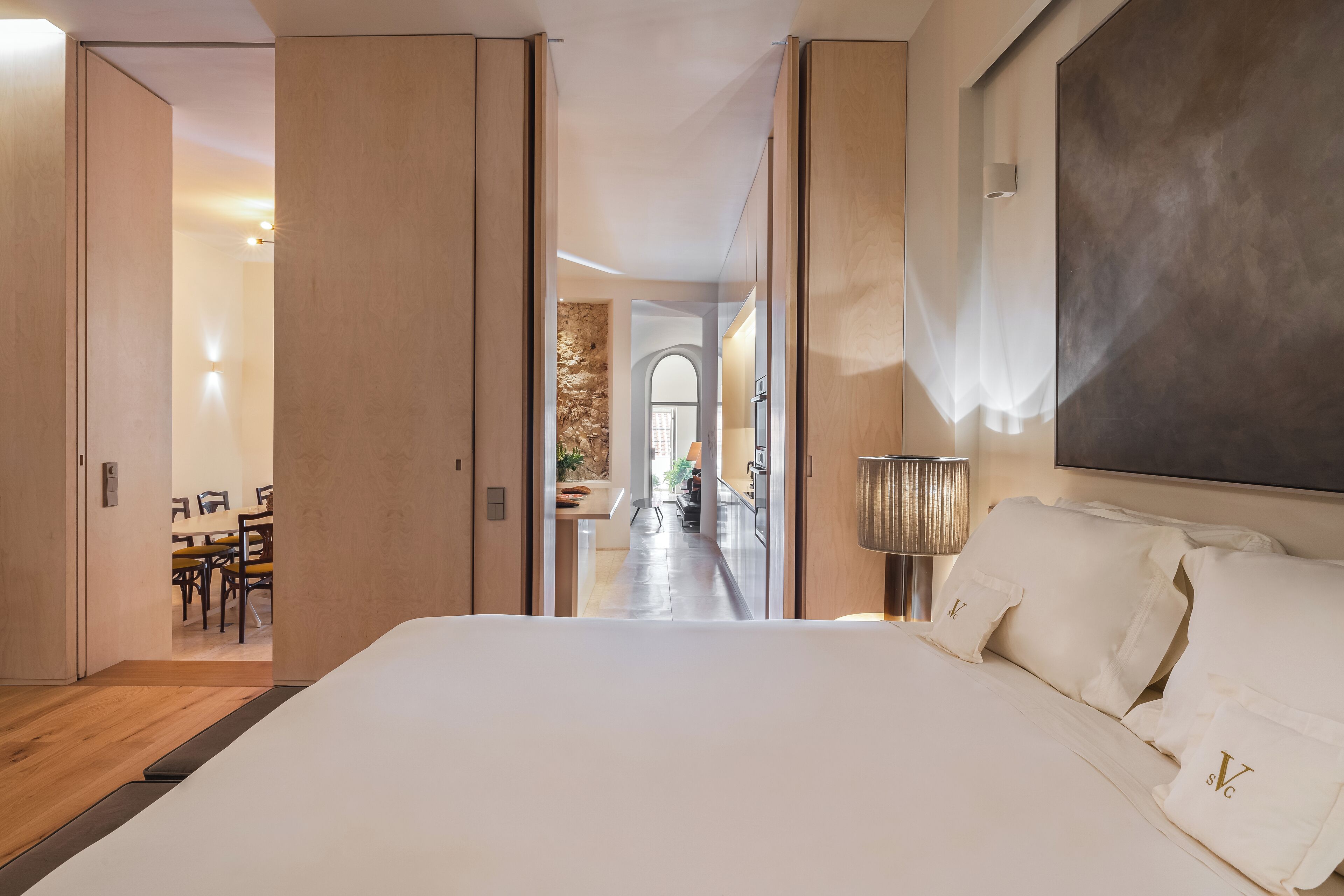 Une chambre d’hôtel avec un grand lit, un bureau et une chaise. On aperçoit, au fond, une salle à manger et une cuisine.