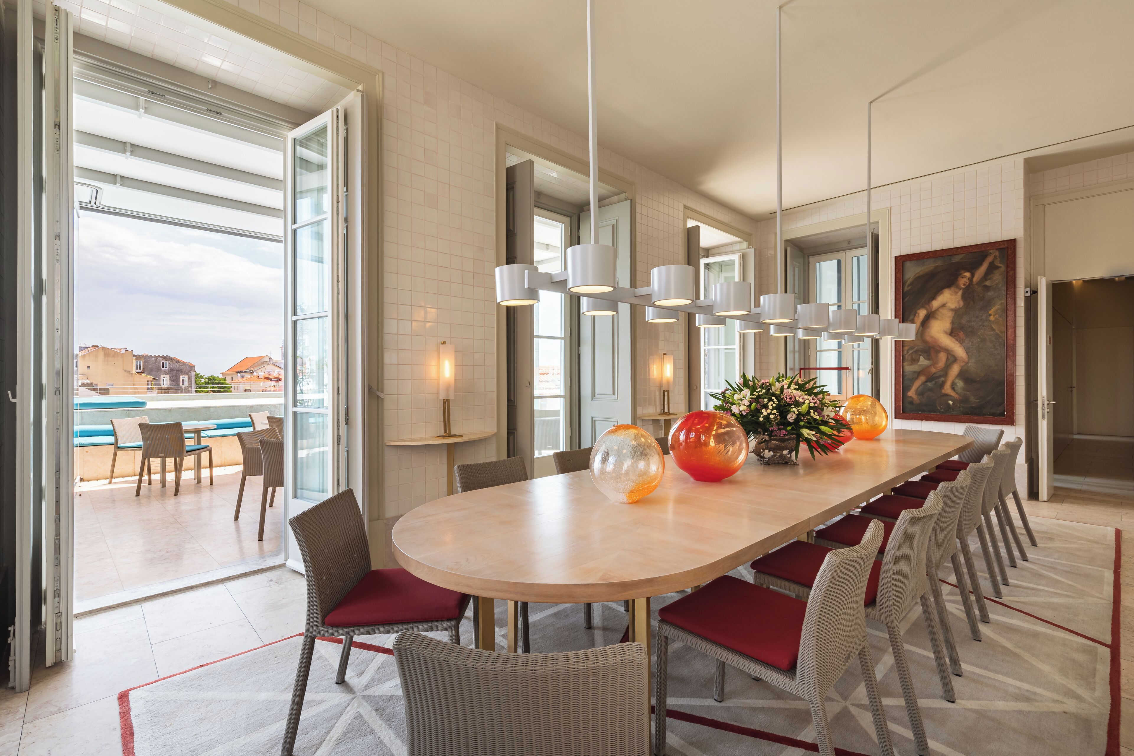 Uma área de jantar moderna com uma mesa oval grande, cadeiras acolchoadas vermelhas e vista para uma piscina.