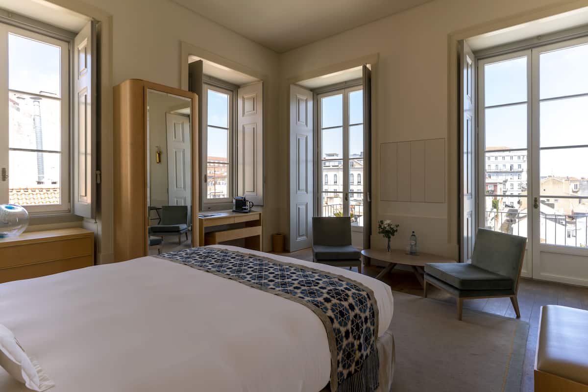Ein Hotelzimmer mit einem großen Bett, einem Schreibtisch, einem Sessel und Blick auf die Stadt durch große Fenster.