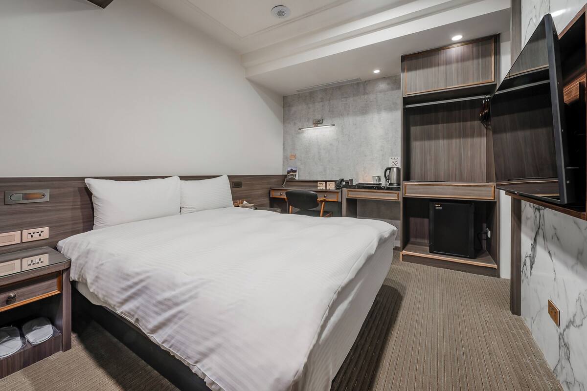 A modern hotel room with a large bed, a desk, a wardrobe, and a compact kitchenette.