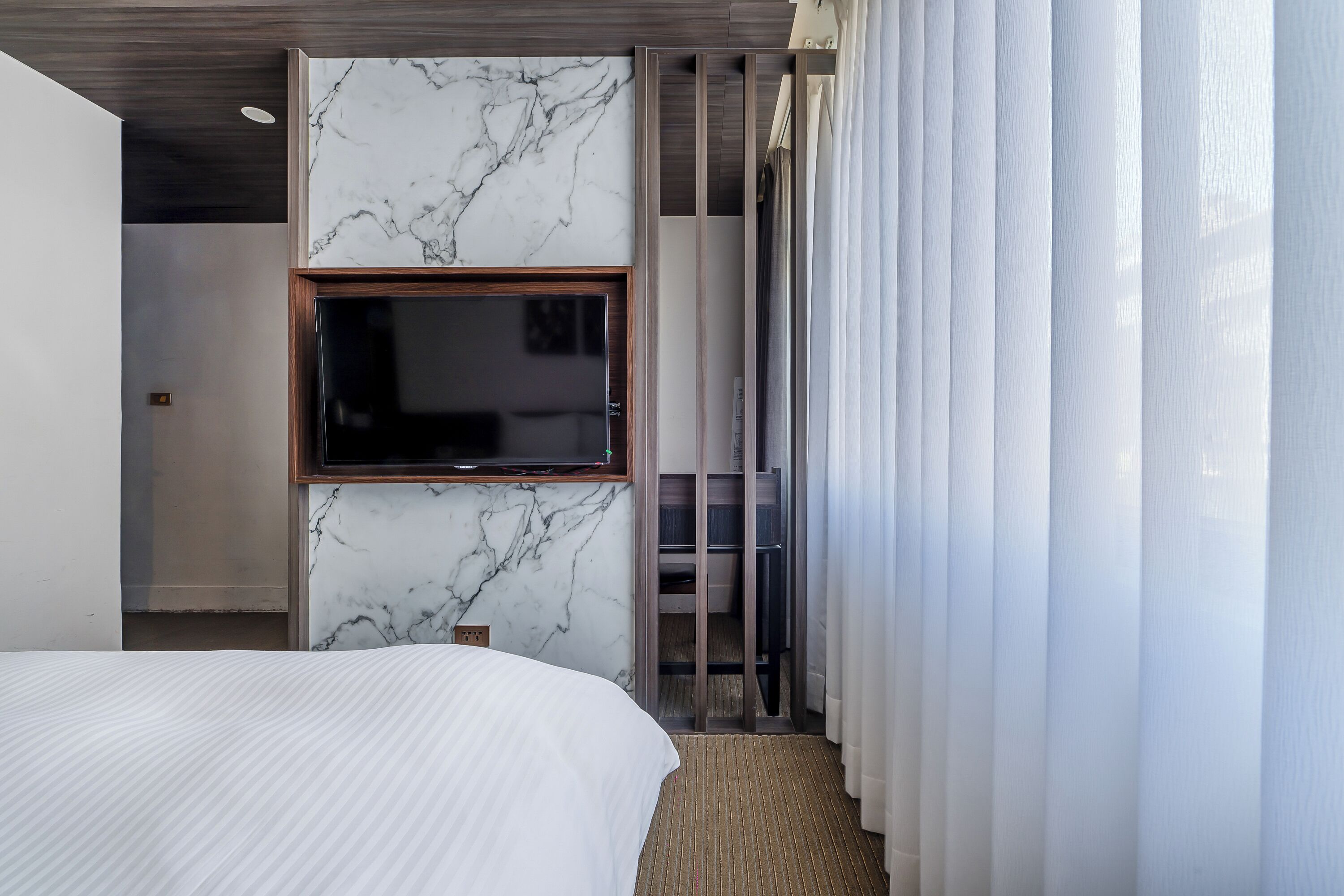 A modern hotel room with a large bed, a flat-screen TV mounted on a marble wall, and a sliding door leading to a balcony.