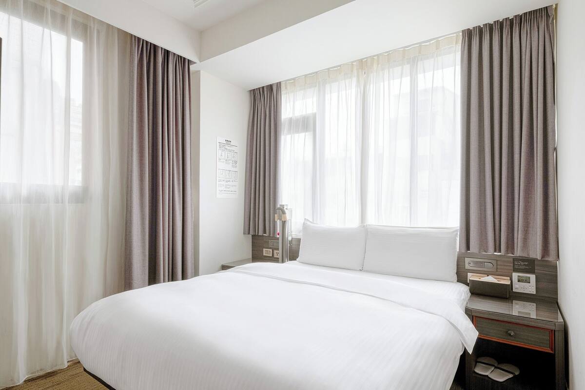 A hotel room with a large bed, two bedside tables, a nightstand, and a window with curtains.