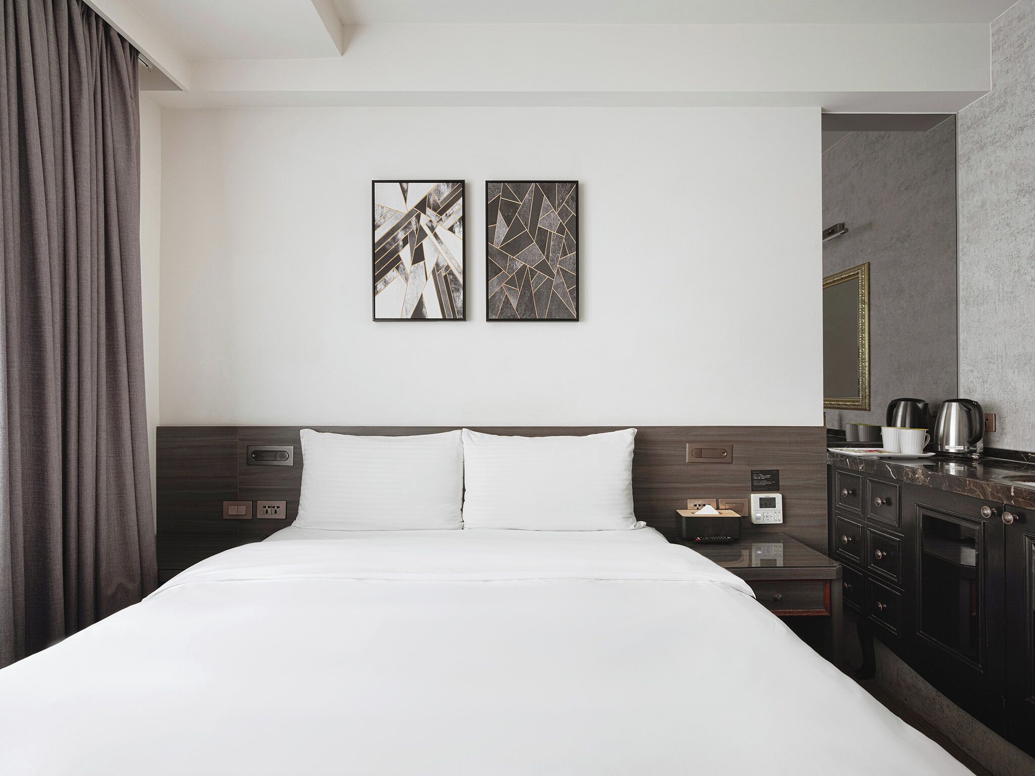 A modern hotel room with a large bed, a nightstand with a lamp, a coffee maker, and a mirror on the wall.