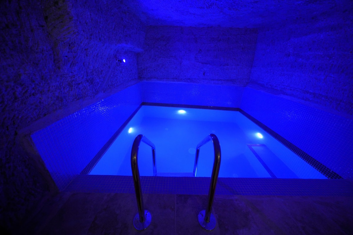 A rectangular hot tub with a tiled interior and metal handrails, set against a stone wall.