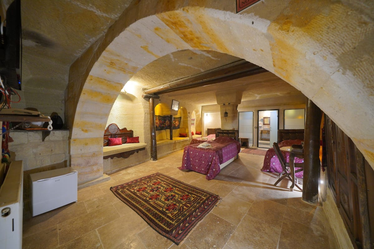 A room with a stone archway, a bed with a purple bedspread, a small table, and a chair.