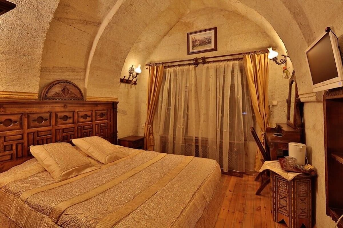 A bedroom with a wooden headboard, a bed with patterned bedding, a wooden nightstand, a television, and a window with curtains.