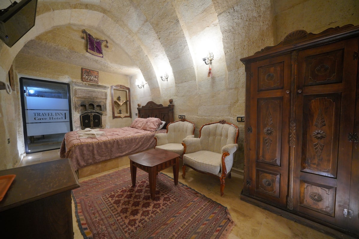 A traditional room with a bed, two armchairs, a wooden cabinet, and a fireplace.