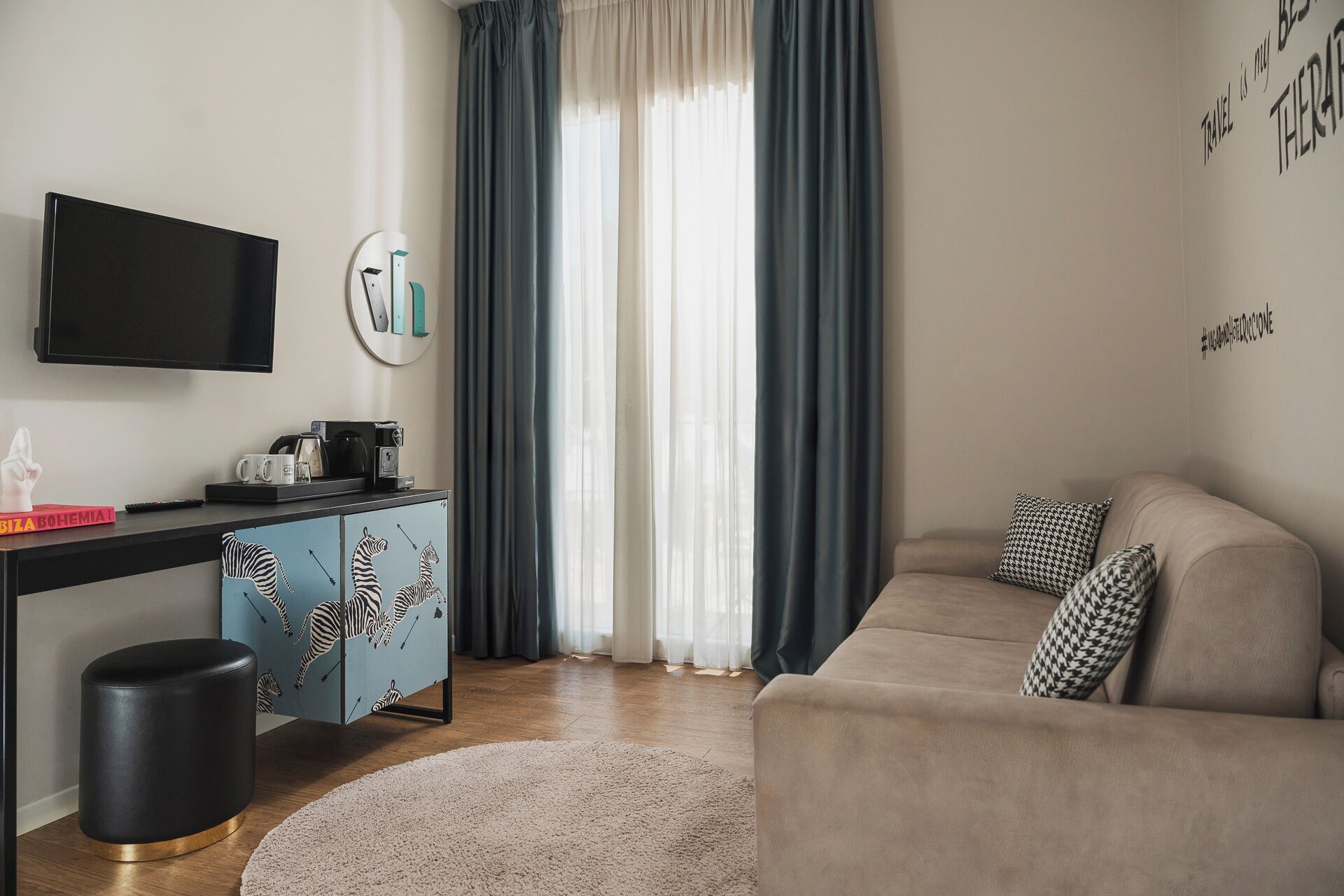 Una sala de estar moderna con un televisor de pantalla plana, un mueble azul con diseños de cebra, un sofá beige con cojines estampados y un ventanal con cortinas oscuras.