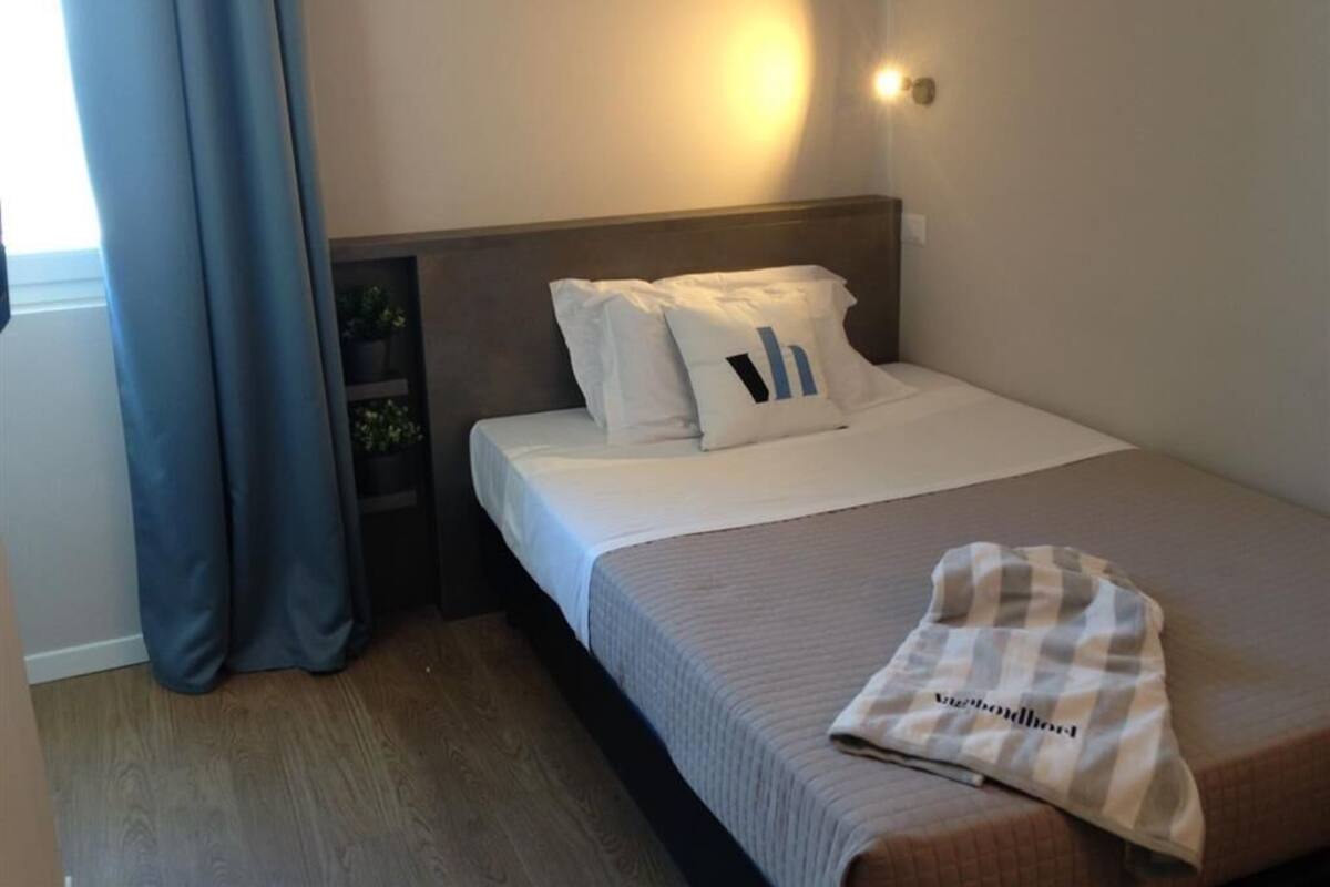 Ein Hotelzimmer mit Bett, Nachttisch, Lampe und einem Fenster mit Vorhängen.