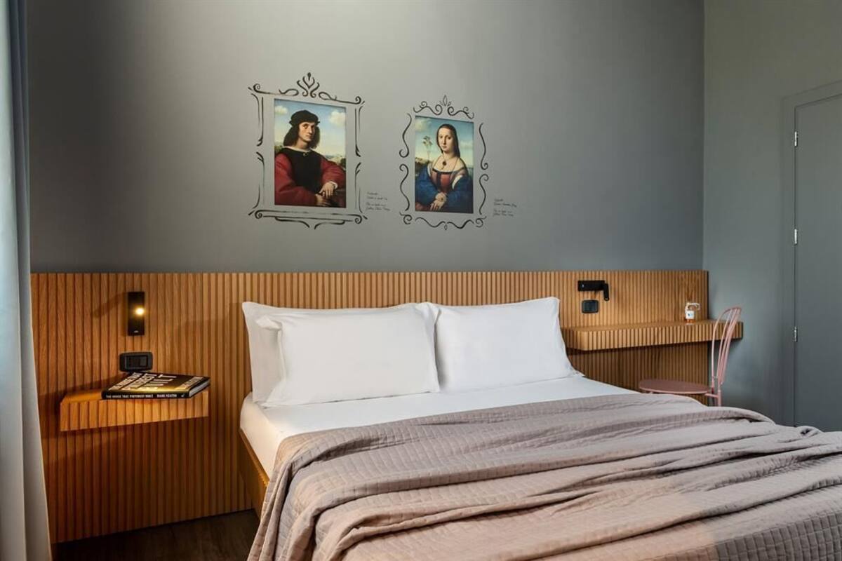 Ein Hotelzimmer mit einem Bett, einem Nachttisch mit einem Buch, einer Wandleuchte und zwei gerahmten Bildern.