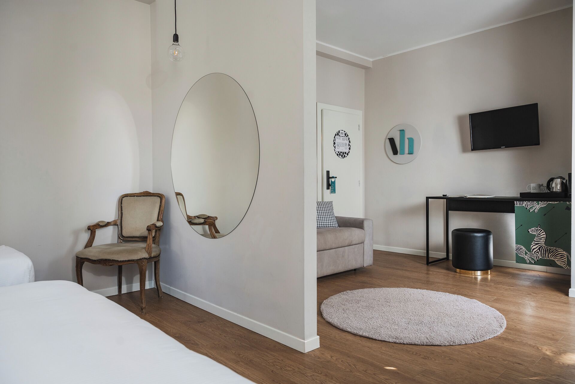 Um quarto com uma cama, uma cadeira de madeira, um espelho redondo, um sofá cinza, um tapete pequeno e redondo, uma escrivaninha com uma televisão e uma lixeira.