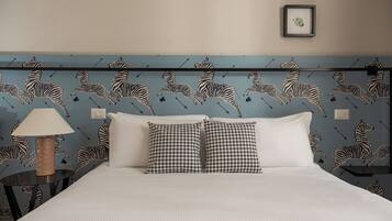 Habitación de hotel con papel tapiz a rayas de cebra, cama con sábanas blancas, dos almohadas estampadas, mesita de noche con lámpara y cuadros en la pared.