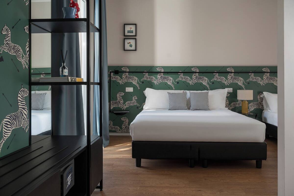 Ein Hotelzimmer mit einem Kopfende im Zebrastreifen-Muster, einem großen Bett mit weißer Bettwäsche, einem Nachttisch mit Lampe sowie einem Kleiderschrank mit Zebradesign.