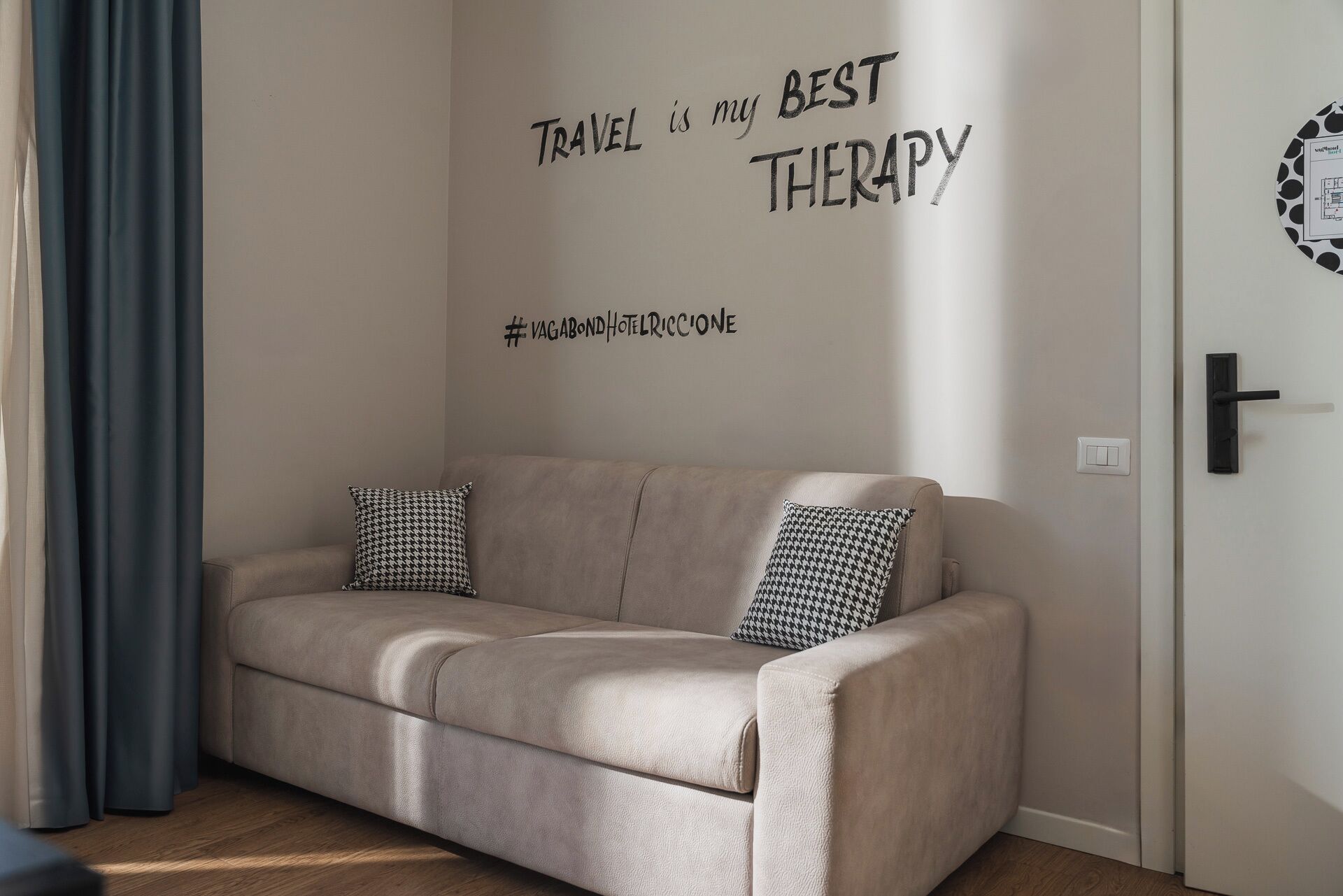 A beige sofa with two patterned cushions, a wall with the text "TRAVEL is my BEST THERAPY" and a hashtag, and a door with a light switch.