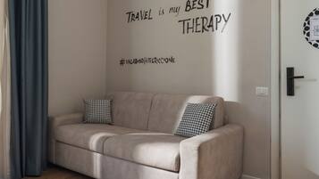 A beige sofa with two patterned cushions, a wall with the text "TRAVEL is my BEST THERAPY" and a hashtag, and a door with a light switch.