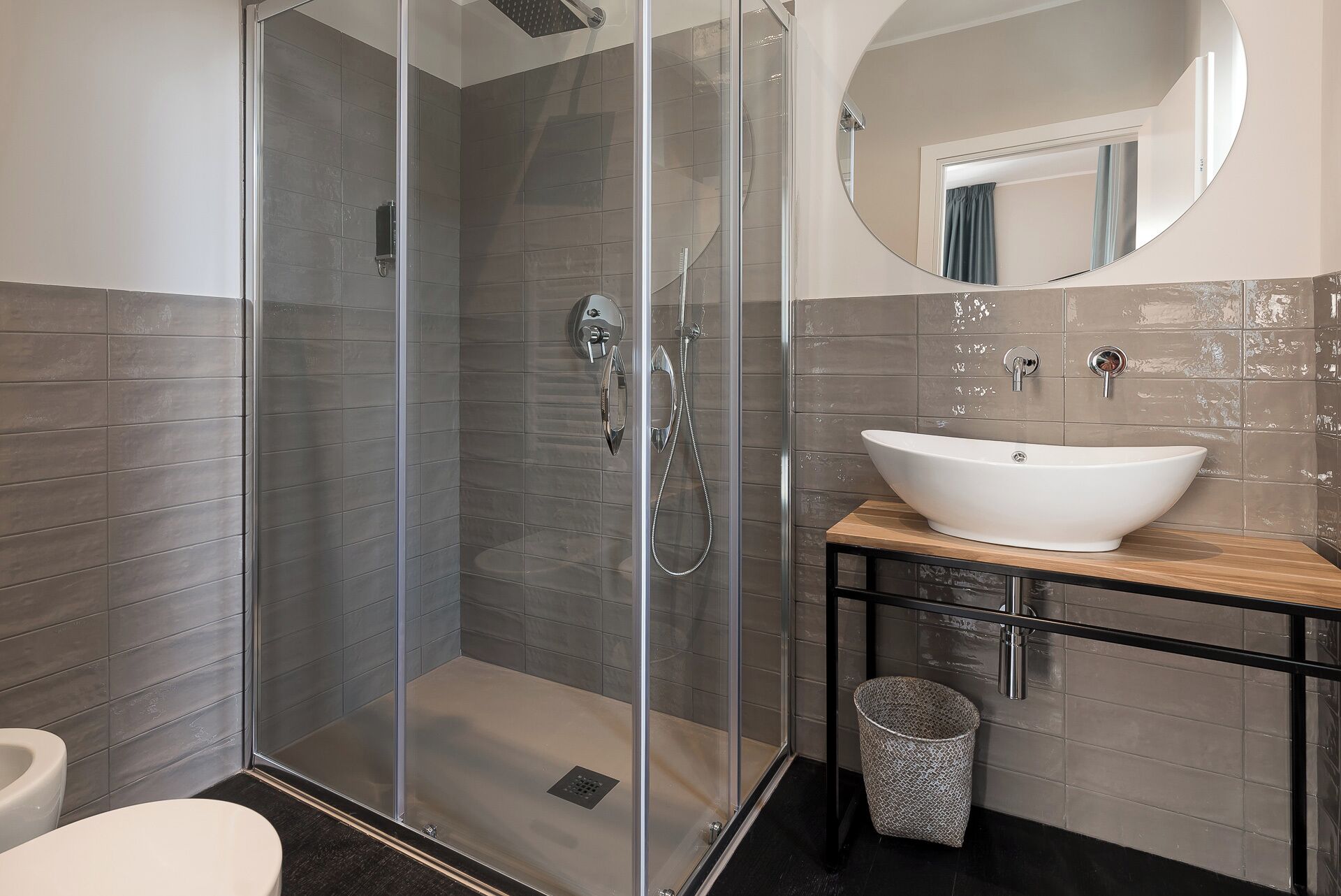 Een moderne badkamer met een douchekabinet van glas, een wit wastafeltje en een ronde spiegel.