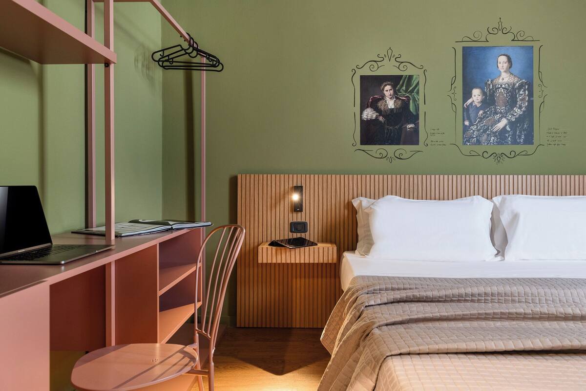 Ein Schlafzimmer mit einem Bett, einem Schreibtisch mit Laptop, einem Sessel und einer Wandleuchte.