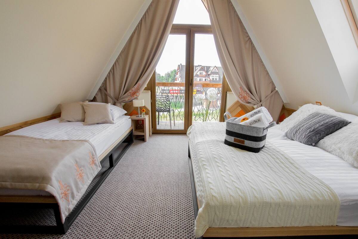 Une chambre avec deux lits, une fenêtre avec des rideaux, une table de chevet avec une lampe, et une vue sur les bâtiments à l’extérieur.