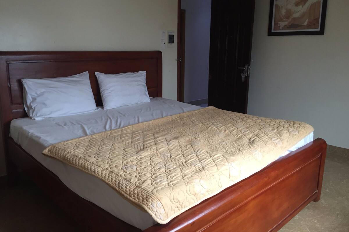 A neatly made bed with a quilted cover and two pillows, a wooden headboard, and a door in the background.