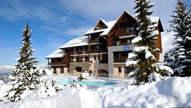 A multi-story lodge with a pool, surrounded by snow-covered trees and mountains. - Hôtel Les Carrettes (Valmeinier)