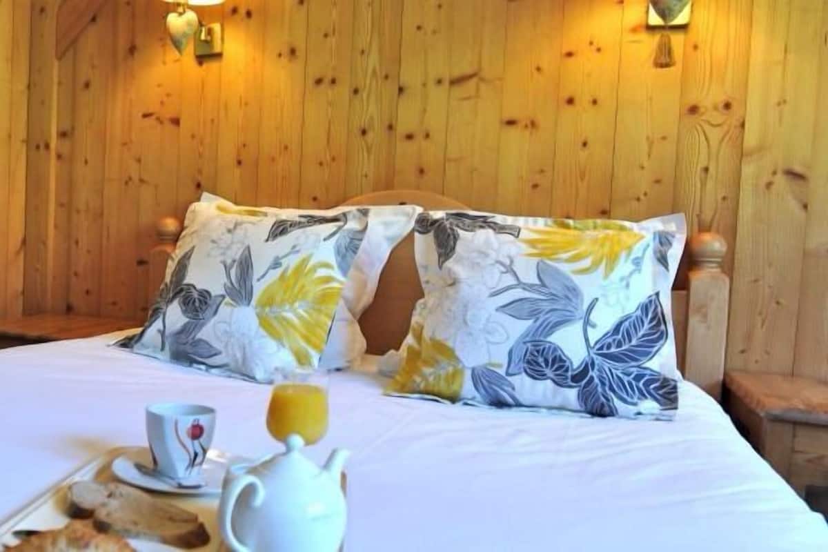 Ein Zimmer mit Holzwänden, einem Bett, Kissen und einem Tablett mit Frühstücksangeboten, beleuchtet von Wandleuchten.