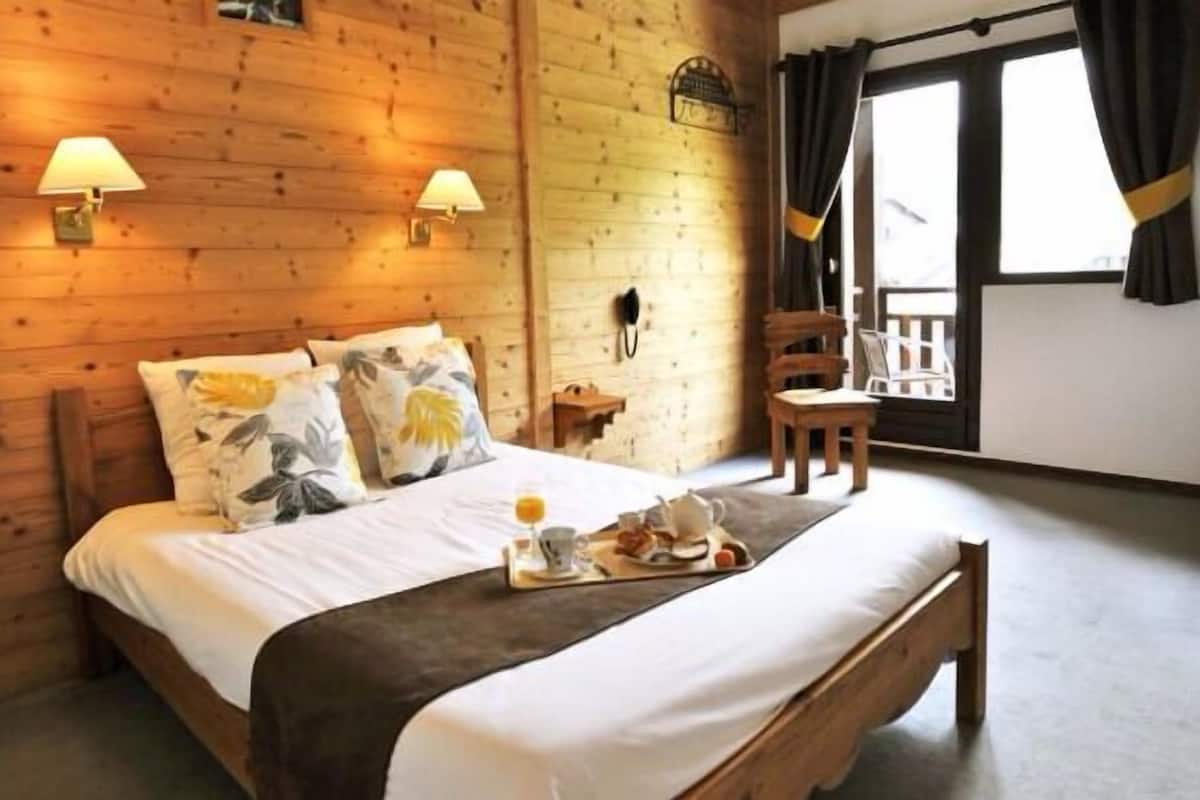 Ein Zimmer aus Holz mit einem Bett, zwei Wandleuchten, einem kleinen Tisch mit Frühstücksgebäck und einem Fenster mit Vorhängen.
