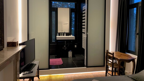 A modern bedroom with a bed, a desk with a laptop, a chair, and a view of a bathroom with a sink and mirror.