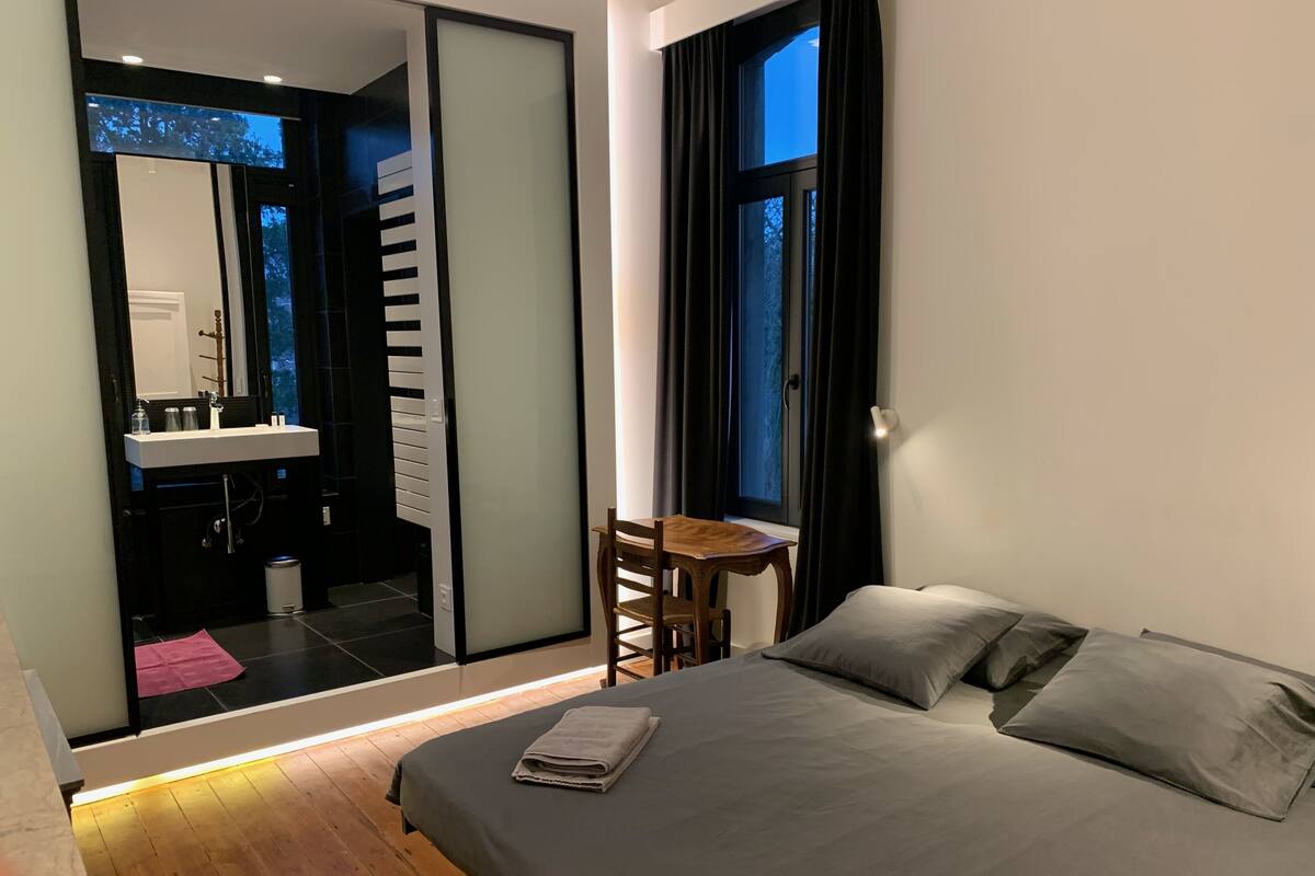 A modern bedroom with a large bed, a bedside table, a chair, and a view of the outdoors through a window.