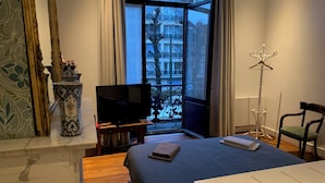 A bedroom with a bed, a television, a window with a view of buildings, a floor lamp, and a chair.