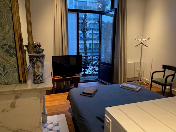 A bedroom with a bed, a television, a window with a view of buildings, a floor lamp, and a chair.