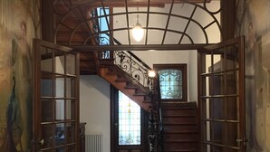 A grand staircase with ornate railings and a chandelier, leading to a room with a large window and a radiator.