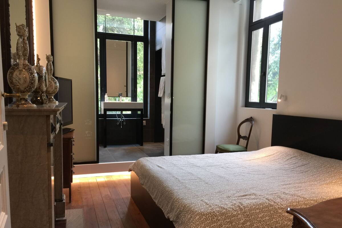 A bedroom with a large bed, a wooden floor, a dresser, a chair, and a view of a bathroom with a sink and mirror.