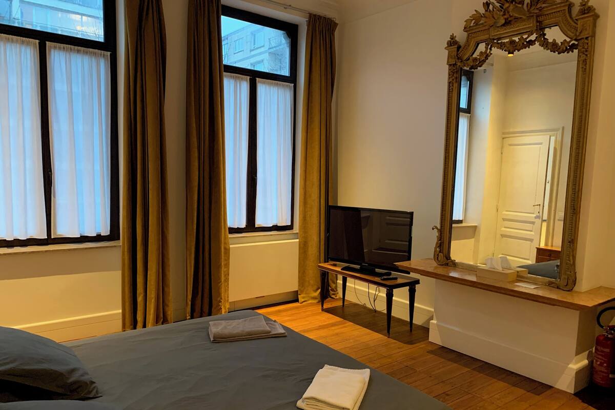 A hotel room with a bed, a television on a stand, a mirror, and a window with curtains.