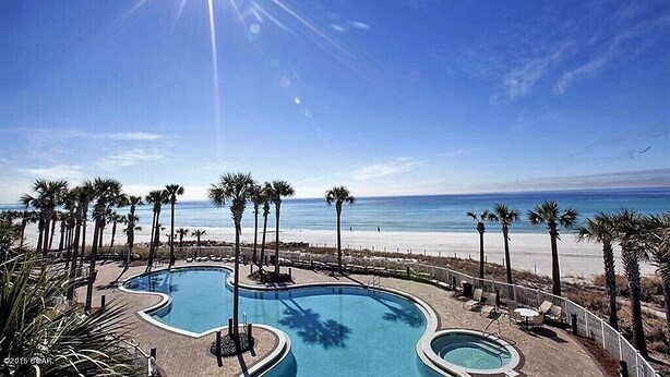A pool area with a curved design, surrounded by palm trees and overlooking a beach. - GRAND PANAMA BEACHFRONT, 1ST FLR DIRECT to BEACH. $250 SpecialSMART TV/FREE WIFI (Panama City Beach)
