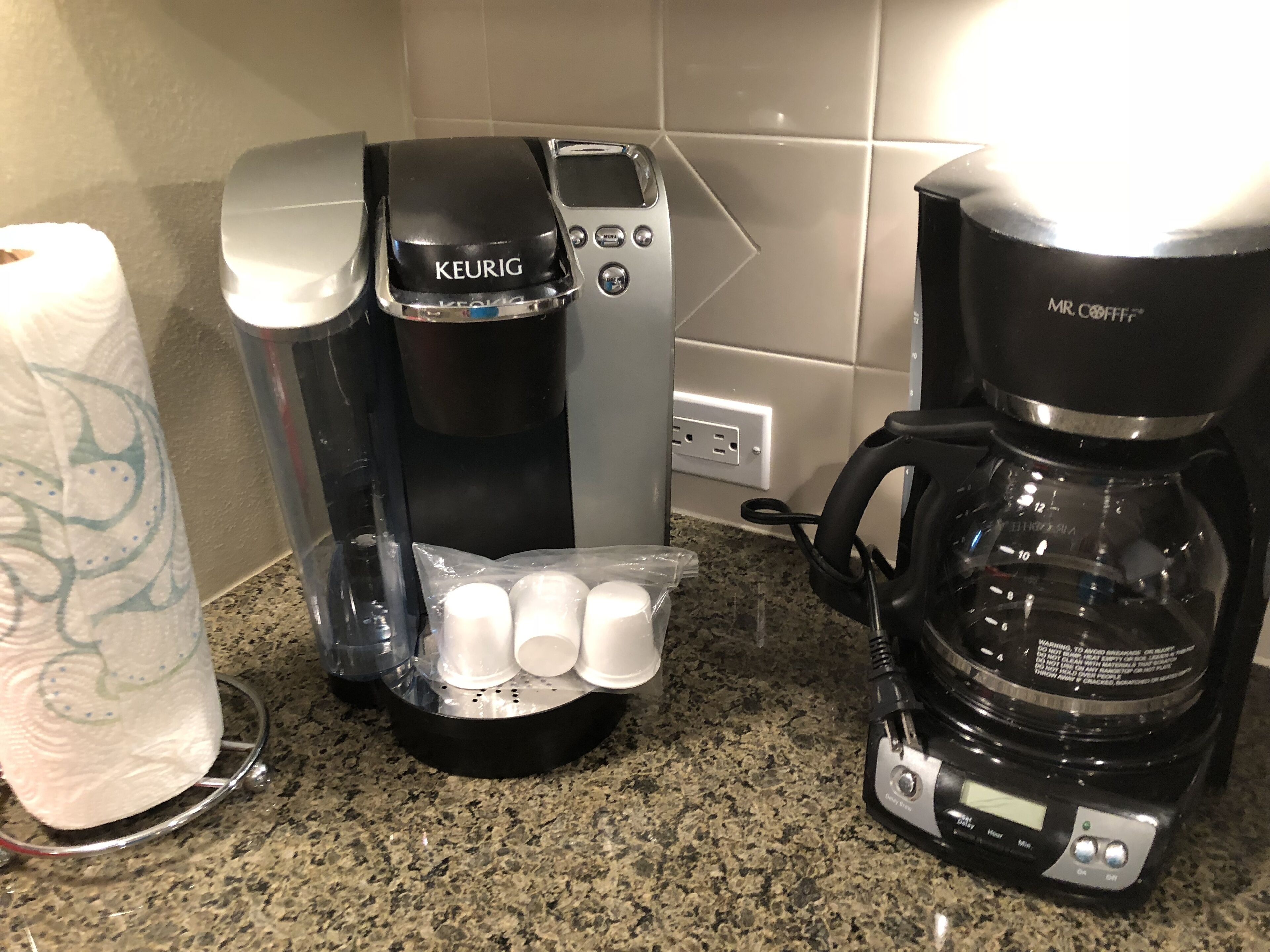 Una cafetera Keurig con un dispensador de toallas de papel y una cafetera Mr. Coffee sobre una encimera de granito.