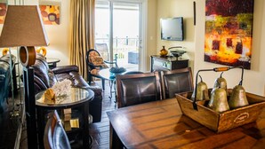 A living room with a leather sofa, a round coffee table, a wooden dining table, and a ceiling fan. There is a TV mounted on the wall and a painting above it. - GRAND PANAMA BEACHFRONT, 1ST FLR DIRECT to BEACH. $250 SpecialSMART TV/FREE WIFI (Panama City Beach)