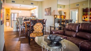 A modern living room with a leather sofa, a glass coffee table, and patterned chairs. A kitchen with white cabinets and a dining area with a table and chairs is visible in the background. - GRAND PANAMA BEACHFRONT, 1ST FLR DIRECT to BEACH. $250 SpecialSMART TV/FREE WIFI (Panama City Beach)