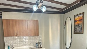 A compact kitchen with white cabinets, a tiled backsplash, and a small refrigerator. There is a dining area with a table and four gray sofas. A ceiling light fixture is visible above the kitchen. - Arifbey Konagi Otel (Gaziantep)