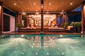 A modern outdoor pool area with a wooden deck, glass doors leading to a living space, and a clear blue pool.
