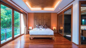 A spacious bedroom with a large bed, wooden paneling on the walls, and a view of lush greenery through large windows. - Villa Malisa (Choeng Thale)