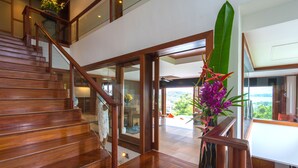 A wooden staircase leading to a second floor with glass railings, a large potted plant with purple flowers, and a view of the outdoors. - Villa Malisa (Choeng Thale)