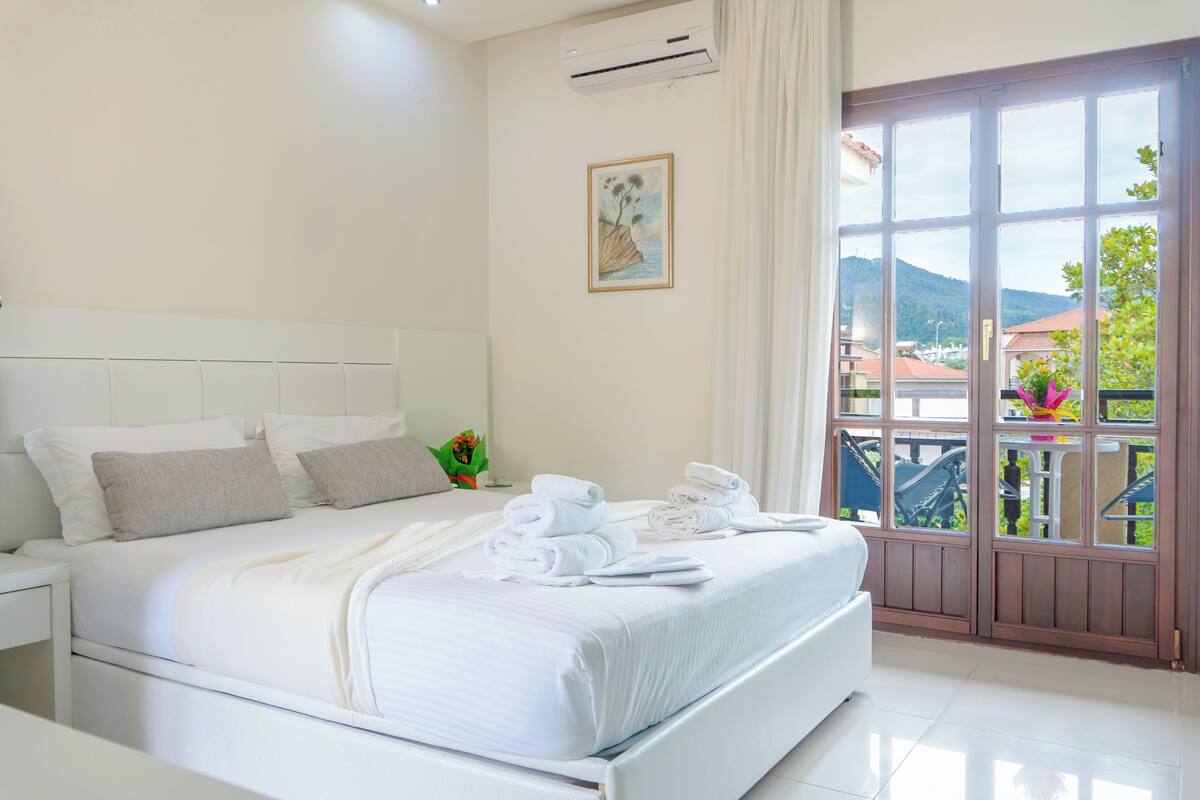 A hotel room with a large bed, white bedding, a nightstand, a window with a view of greenery and buildings, and a painting on the wall.